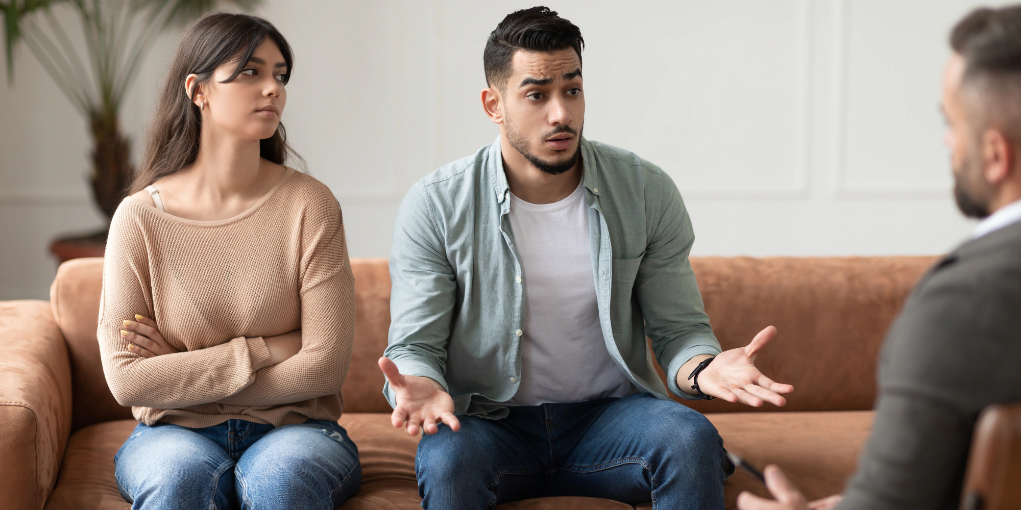 A wife sits with her arms crossed looking away while her husband gestures mid-explanation during a marriage counseling session — the disconnection that porn addiction creates between spouses